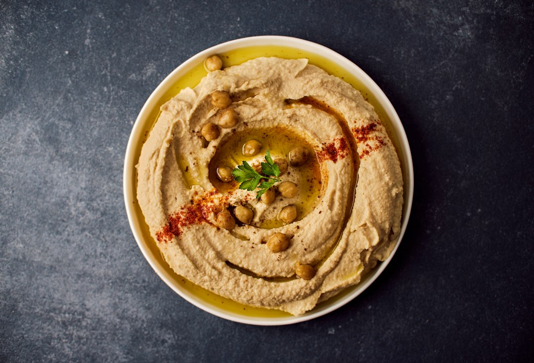 A bowl of creamy hummus drizzled with olive oil, topped with whole chickpeas, a sprig of parsley, and sprinkled with paprika.